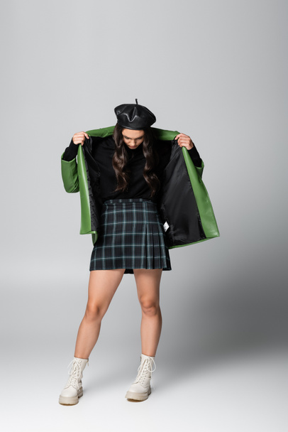 full length of young stylish woman in beret and plaid skirt adjusting green leather jacket on grey - 写真・画像