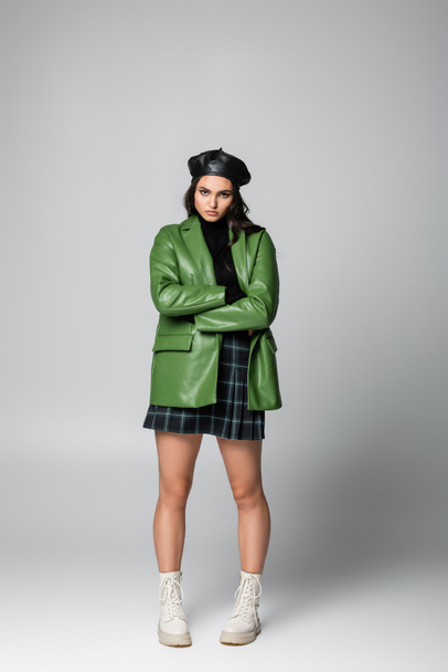 полная длина молодой стильной женщины в берете, клетчатой юбке и зеленой кожаной куртке, позирующей со скрещенными руками на сером - Фото, изображение