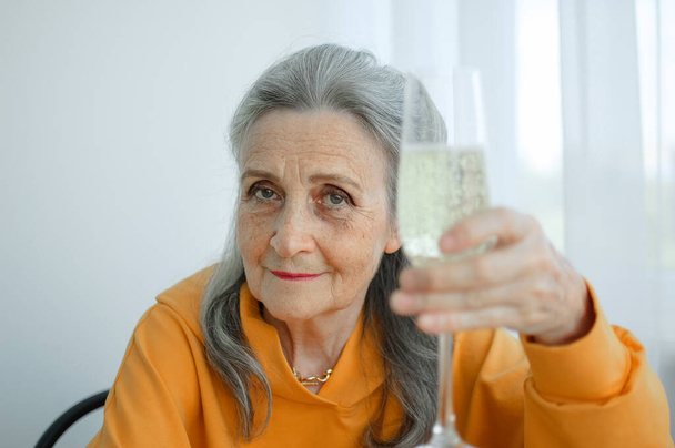 Beautiful old grandmother with grey hair and face with wrinkles sitting at the table at home on window background with glass of champagne, mothers day, happy retirement - Photo, Image