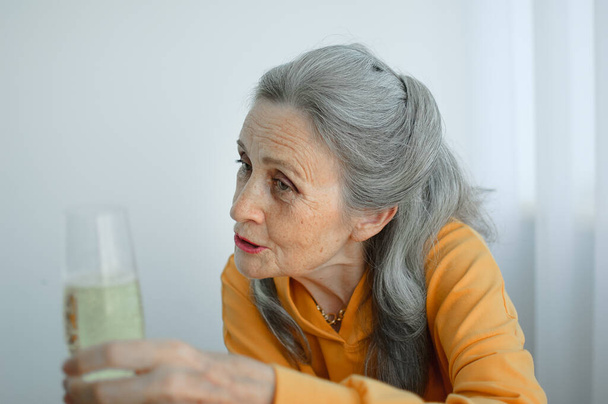 Schöne alte Großmutter mit grauen Haaren und faltenbehaftetem Gesicht, die zu Hause am Tisch sitzt, auf Fensterhintergrund mit einem Glas Champagner, Muttertag, glücklicher Ruhestand - Foto, Bild