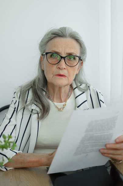 Senior grijs harige zakenvrouw in gestreepte jas met bril werkt met documenten hier kantoor zitten aan het bureau. Gelukkige pensionering, werkgelegenheid en arbeid - Foto, afbeelding