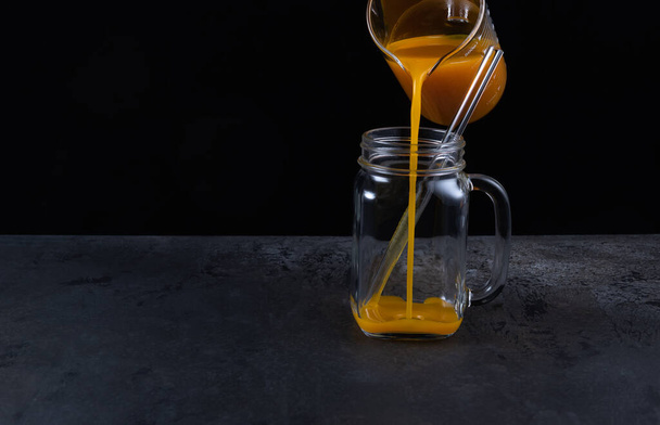 Pouring bright orange non-alcoholic pumpkin mocktail in square glass jar with glass straw on black textured table surface. Horizontal background with copy space. Healthy food cooking. Selective focus. - Photo, image