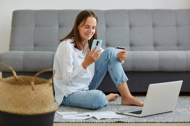 Smiling woman wearing white shirt and jeans sitting near sofa, holding smart phone and credit card in hands, making online payments, looking at phone screen. - 写真・画像
