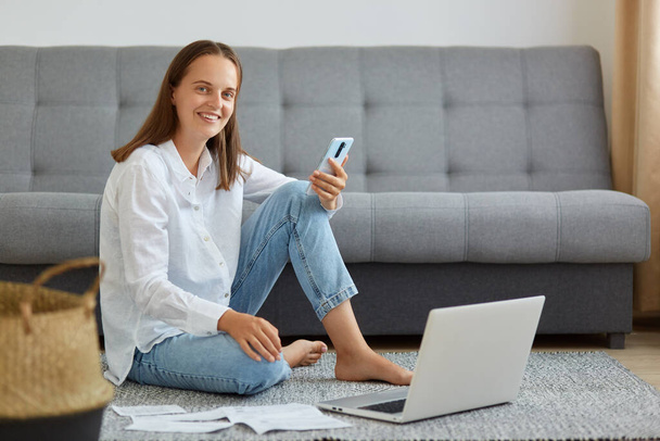 Positive female wearing white shirt and jeans sitting on floor in living room, holding cell phone in hands, posing near laptop, looking at camera with toothy smile, freelancer working online. - Фото, зображення