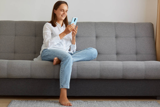 Young adult attractive female wearing white shirt and jeans sitting on the sofa in living room and using smart phone, blogger writing post for social networks. - Zdjęcie, obraz