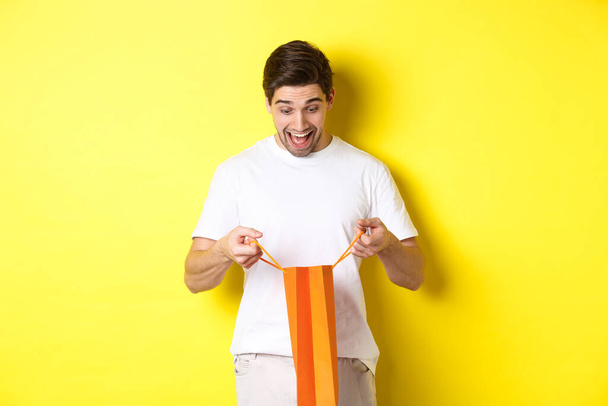 Возбужденный парень открытый мешок с подарком, глядя внутрь с изумлением и счастливым лицом, стоя на желтом фоне - Фото, изображение