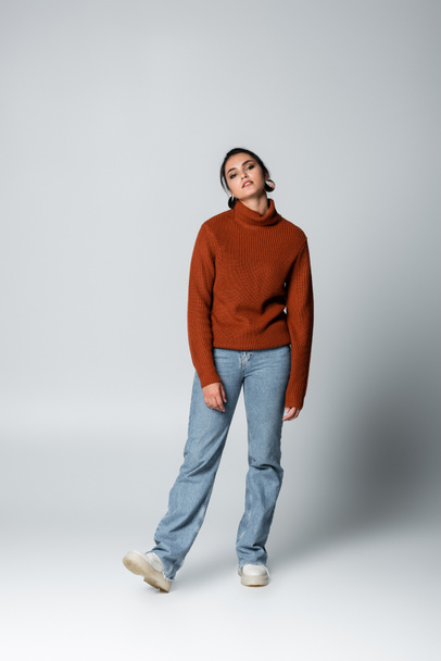full length of young woman in sweater and denim jeans on grey - Fotoğraf, Görsel