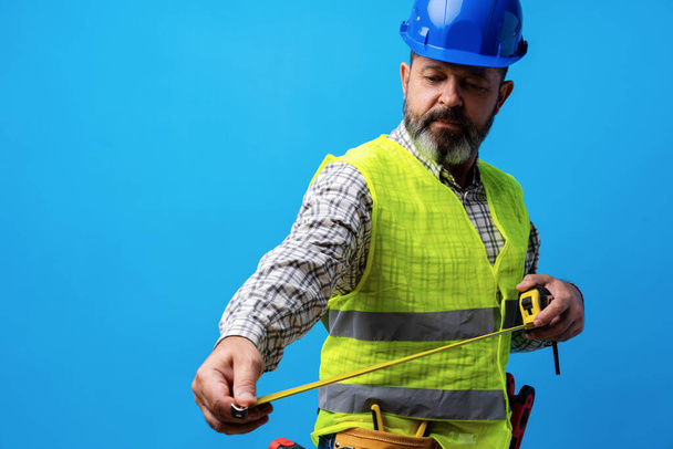 Male builder holding measuring roulette against blue background - Foto, immagini