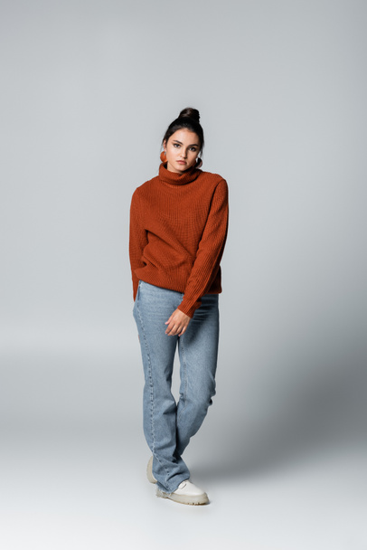 full length of young woman in sweater and blue jeans posing on grey - Фото, изображение