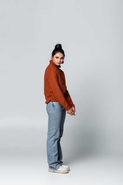 full length of young model in sweater and denim jeans posing on grey - Foto, afbeelding