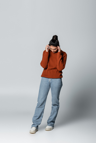 volledige lengte van jonge vrouw in trui en spijkerbroek kijken neer op grijs - Foto, afbeelding
