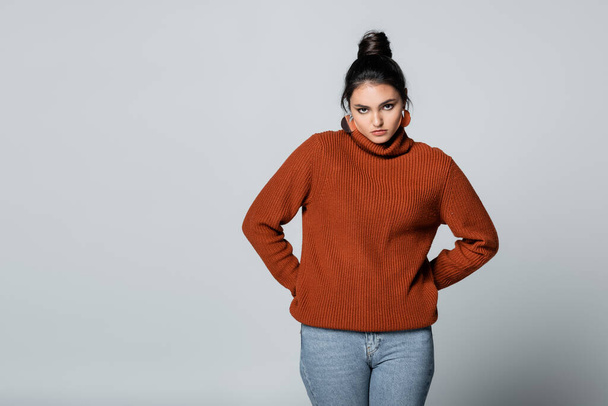 young woman in sweater and denim jeans posing with hands on hips isolated on grey - Foto, Imagen