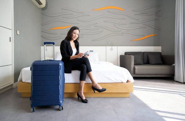 Mujer de negocios asiática en traje negro sentada con tableta en la mano. Su equipaje colocado al lado de la cama. Ambiente de trabajo al aire libre durante las vacaciones. - Foto, Imagen