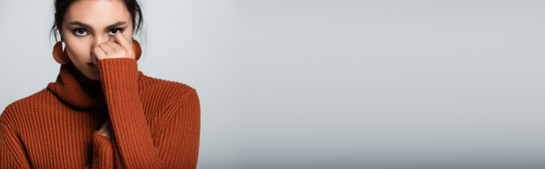 young woman in warm sweater looking at camera while covering face isolated on grey, banner - Foto, imagen