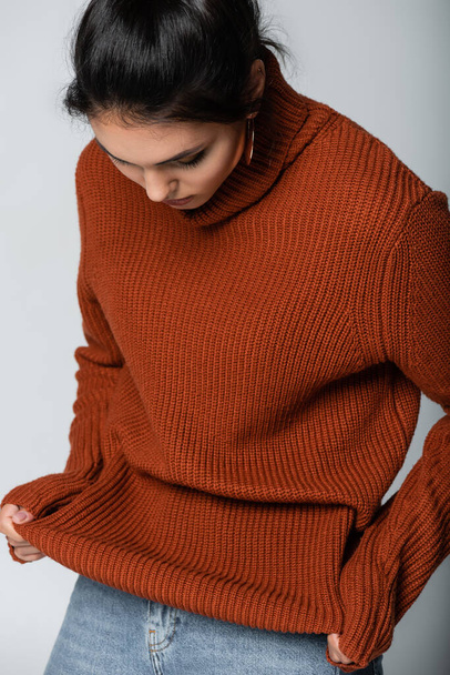 mladá žena v náušnici při pohledu na pletený svetr izolované na šedé - Fotografie, Obrázek