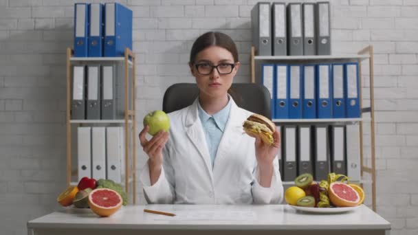 Professionista nutrizionista signora mostrando mela verde e hamburger spazzatura alla fotocamera, confrontando nutrizione sana e malsana - Filmati, video