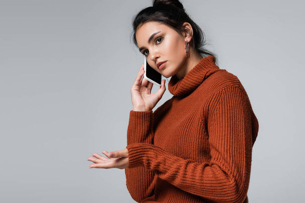 young woman in knitted sweater talking on smartphone while gesturing isolated on grey - Fotó, kép