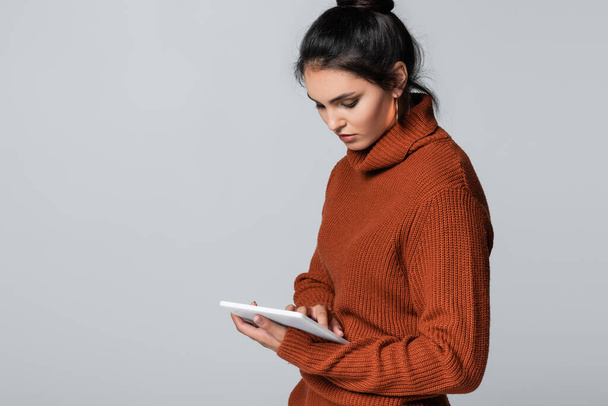 グレーに隔離されたデジタルタブレットを使ってニットセーターを着た若い女性  - 写真・画像