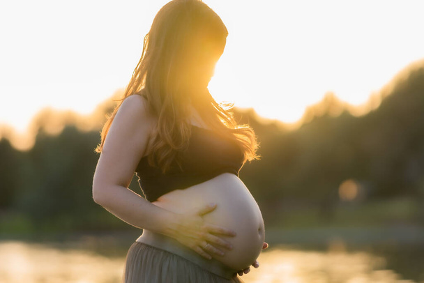 Sonnenuntergang erhellt eine schwangere Frau, als sie ihren Bauch im Freien berührt - Foto, Bild