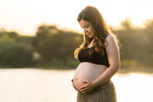 Pregnant woman looking and touching her belly while standing in a park - Fotografie, Obrázek