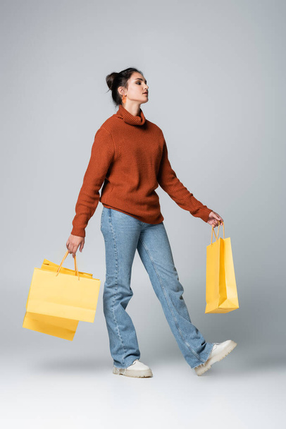 full length of young model in sweater and jeans holding yellow shopping bags on grey  - Фото, зображення