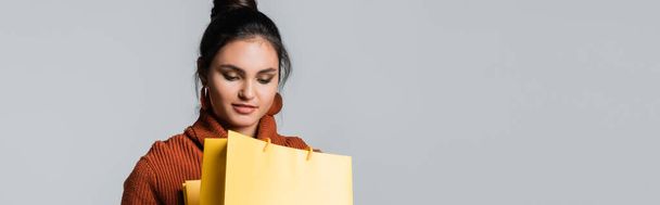 young woman in sweater looking at yellow shopping bag isolated on grey, banner - Fotoğraf, Görsel