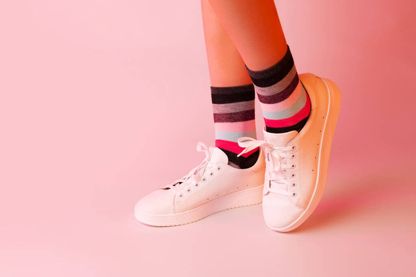 Moda - scarpe da ginnastica in pelle bianca, calze a righe colorate e gambe da ragazza. Calzature su sfondo rosa pastello. Layout con spazio libero di copia. - Foto, immagini
