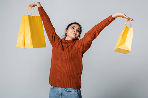 pleased young woman in sweater and jeans holding yellow shopping bags isolated on grey  - Photo, Image