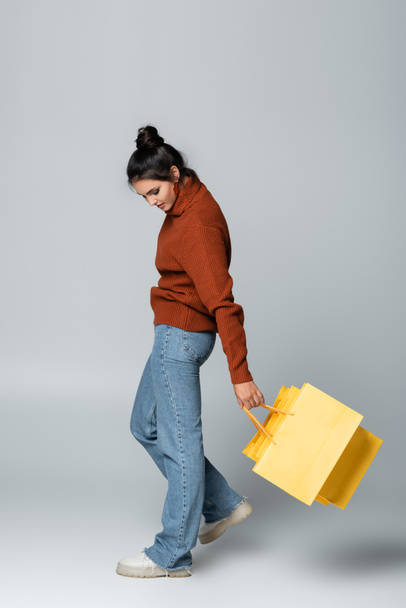 πλήρες μήκος της νεαρής γυναίκας σε πουλόβερ κρατώντας κίτρινες σακούλες για ψώνια και το περπάτημα σε γκρι  - Φωτογραφία, εικόνα