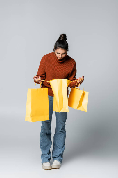 πλήρες μήκος της νεαρής γυναίκας σε πουλόβερ και τζιν κρατώντας κίτρινες σακούλες για ψώνια σε γκρι  - Φωτογραφία, εικόνα
