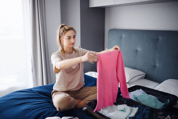 Portrait of young woman packing clothes into suitcase indoors at home, holiday concept. - Фото, изображение