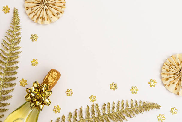 Festive white background with gold decoration , bottle of sparkling wine and paper christmas tree decorations , glittering snowflakes and shiny golden fern leaves, flat lay, top view, copy space - Foto, Imagem