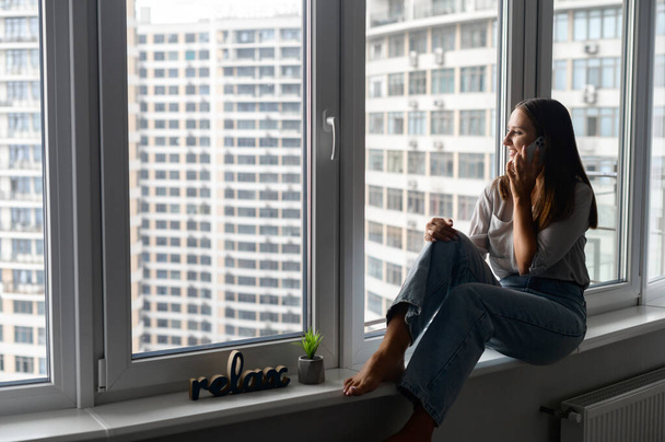 Charming woman talking on the smartphone sitting on the window sill at home - Φωτογραφία, εικόνα