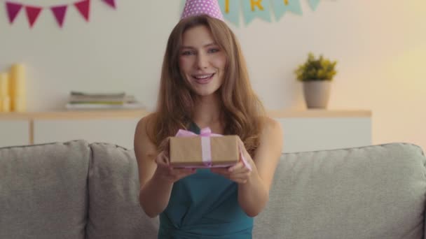 Linda hermosa dama joven en paty cap ofreciendo caja de regalo a la cámara y diciendo feliz cumpleaños, sentado en casa decorada - Imágenes, Vídeo