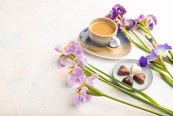 Beker cioffee met chocolade snoepjes en lila iris bloemen op witte betonnen achtergrond. zijaanzicht, kopieerruimte, stilleven. Ontbijt, ochtend, lente concept. - Foto, afbeelding