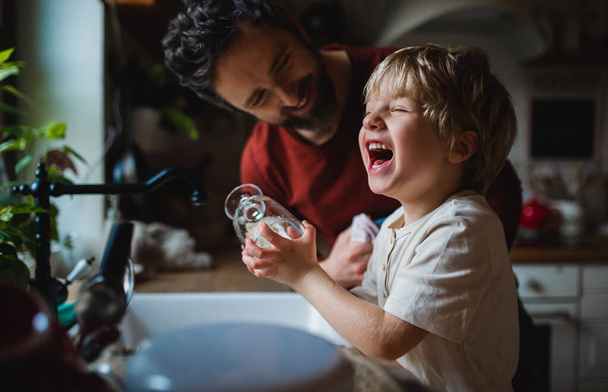 Small laughing boy with father having fun when washing dishes indoors at home, daily chores concept. - Foto, Bild
