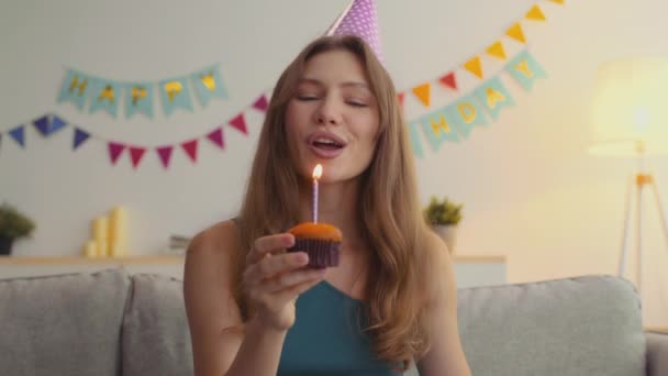 Online birthday party. Webcam portrait of happy young woman wearing party cap blowing up candle on cupcake and dancing - Footage, Video