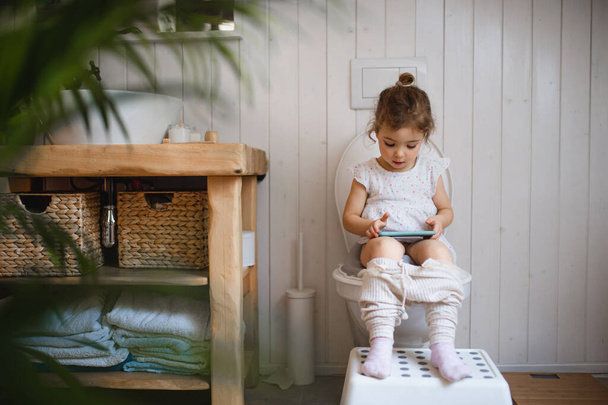 Portrait of cute small sitting on toilet indoors at home, using smartphone. - Valokuva, kuva