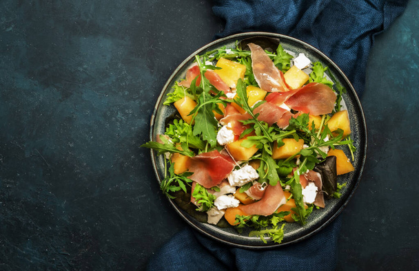 Fresh salad with cantaloupe melon, prosciutto, soft cheese and arugula on blue background, top view, copy space - Photo, Image