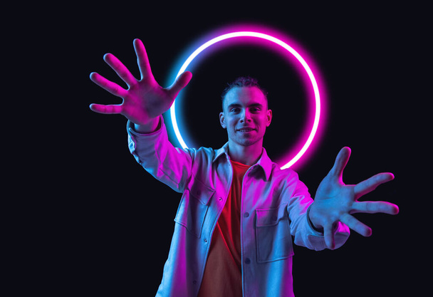 Close-up young happy man gesturing isolated on dark studio background in multicolored neon light with geometric luminescent shape cirlce - 写真・画像