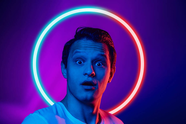 Shocked, scared. Young mans portrait isolated on blue studio background in multicolored neon light with geometric luminescent shape cirlce. Concept of human emotions, facial expression, ad, fashion. - 写真・画像