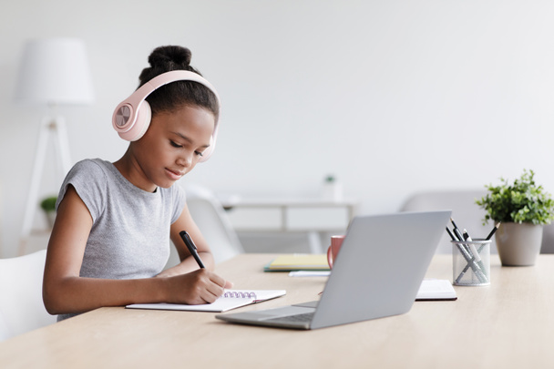 Keskittynyt nuori musta tyttö kuulokkeissa, tekee muistiinpanoja ja tutkimuksia kannettavassa tietokoneessa - Valokuva, kuva