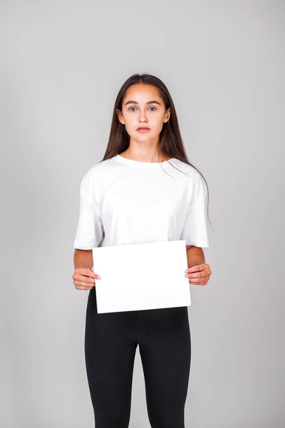 Young beautiful girl holding a blank sheet of paper in her hands, isolated on gray background - Foto, Imagem