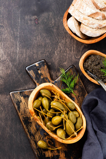 Canned capers in wooden dish, tapenade and bread on trendy dark wooden old background. Top view - 写真・画像