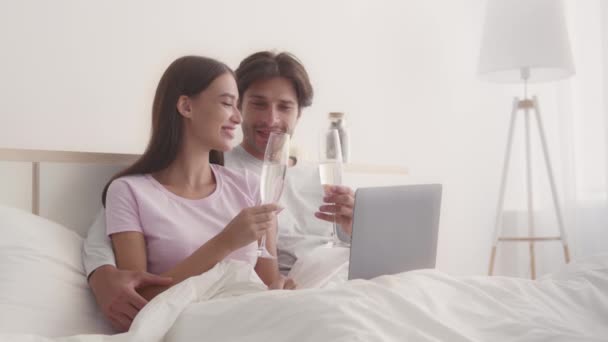 Joven pareja casada tintineando con copas de champán, disfrutando de romántica mañana en la cama con portátil, cámara lenta - Imágenes, Vídeo