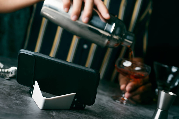 a young caucasian man, wearing a striped shirt, is preparing a cocktail in a metal shaker, following a tutorial in his smartphone - Фото, изображение