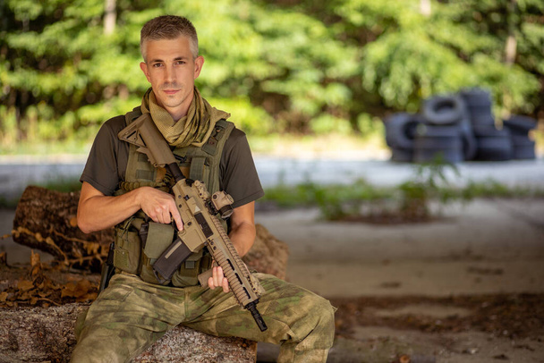 Un uomo seduto con un fucile d'assalto airsoft in uniforme militare nell'hangar di cemento - Foto, immagini