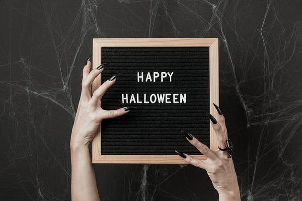 Happy Halloween on letter board in hands with long black nails and spider ring on black background with cobwebs. Halloween holiday concept - Foto, imagen