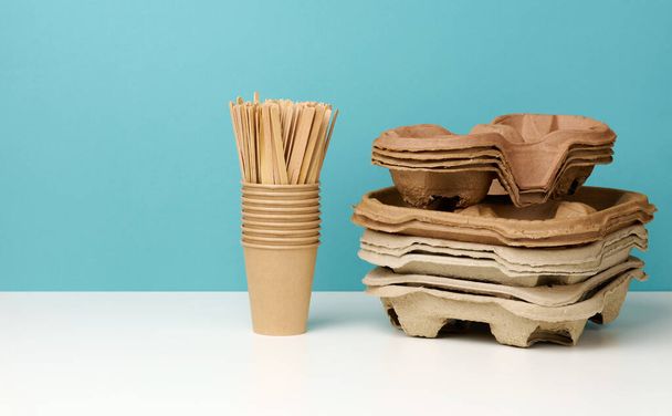 una pila de vasos de papel desechables de color marrón y un soporte sobre una mesa blanca, fondo azul. Utensilios para bebidas para llevar, cero residuos - Foto, imagen
