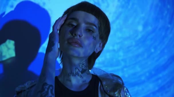 Νεαρό μοντέλο με τατουάζ που αγγίζει το πρόσωπο σε αφηρημένο μπλε φόντο  - Πλάνα, βίντεο